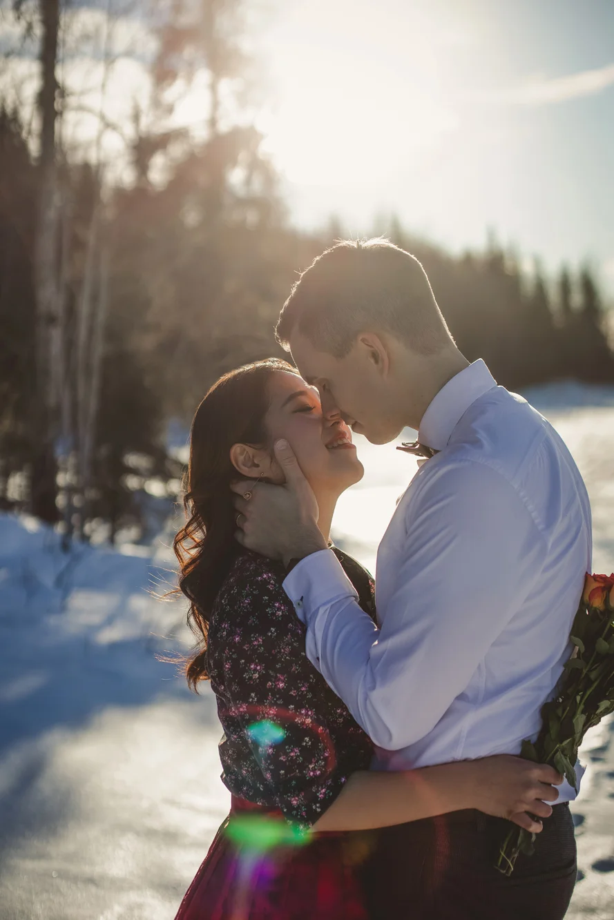 A couple in love stands close together in a snowy outdoor setting during sunset, with the man holding a bouquet of roses and gently touching the woman's face.