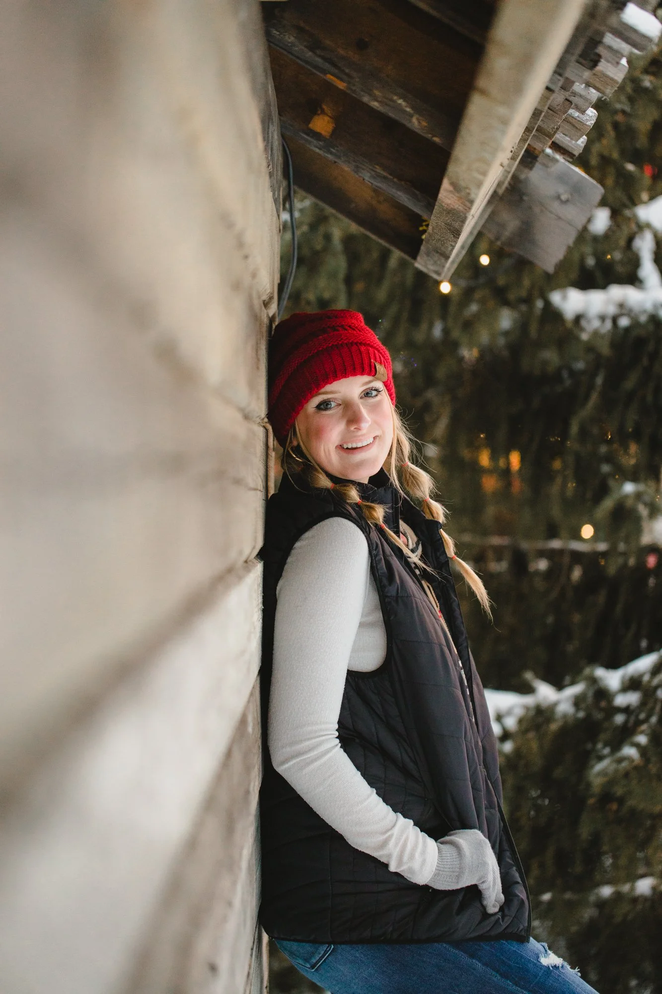 A young woman with blonde hair in braids, wearing a red knit hat, white long sleeve shirt, black puffer vest, and ripped blue jeans, is smiling and leaning against a wooden wall outdoors, with trees and snow in the background during winter.