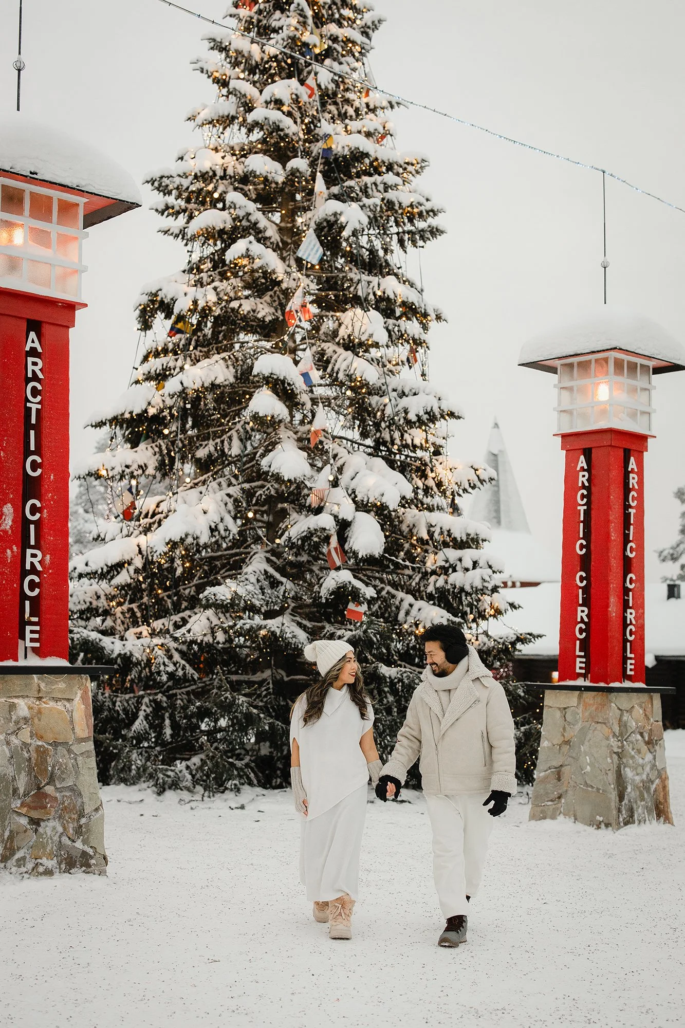 A couple dressed in white winter attire walking hand in hand in front of a snow-covered Christmas tree decorated with lights and ornaments, at a snowy outdoor location with arching red signposts labeled 'ARCTIC CIRCLE' on stone bases.