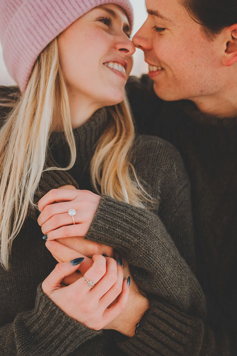 Close-up of a smiling couple with noses touching, embracing, woman wearing a pink knit hat and diamond ring, woman with dark nail polish and rings, man with short dark hair, both wearing dark sweaters.