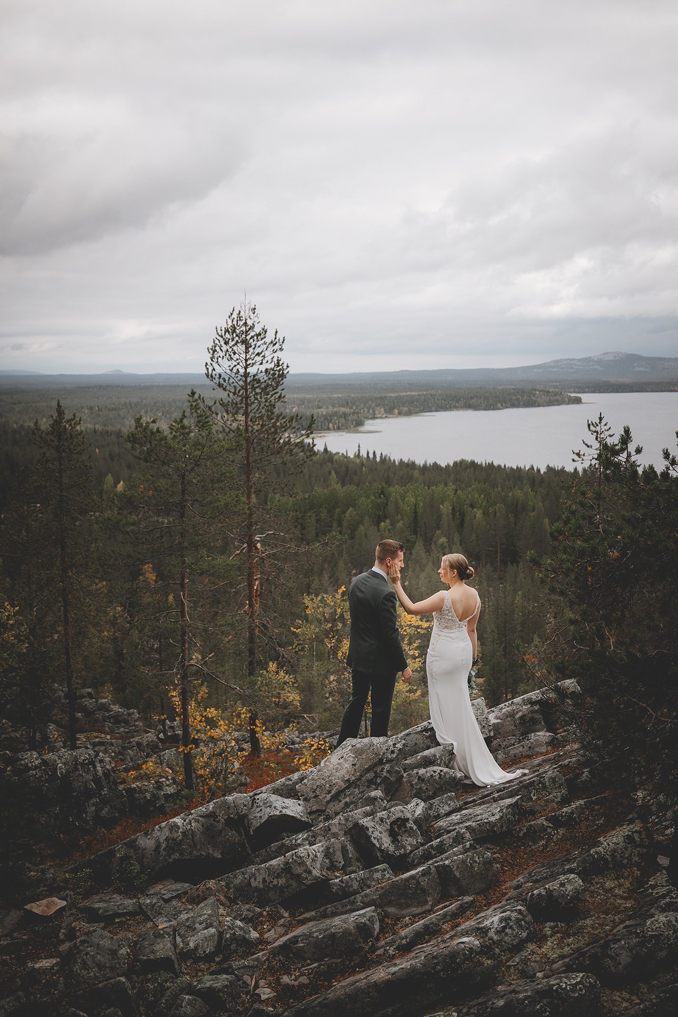 A bride and groom holding hands and gazing into each other's eyes on a rocky ledge overlooking a forested landscape with a lake and hills in the background under an overcast sky. Wedding photographer.