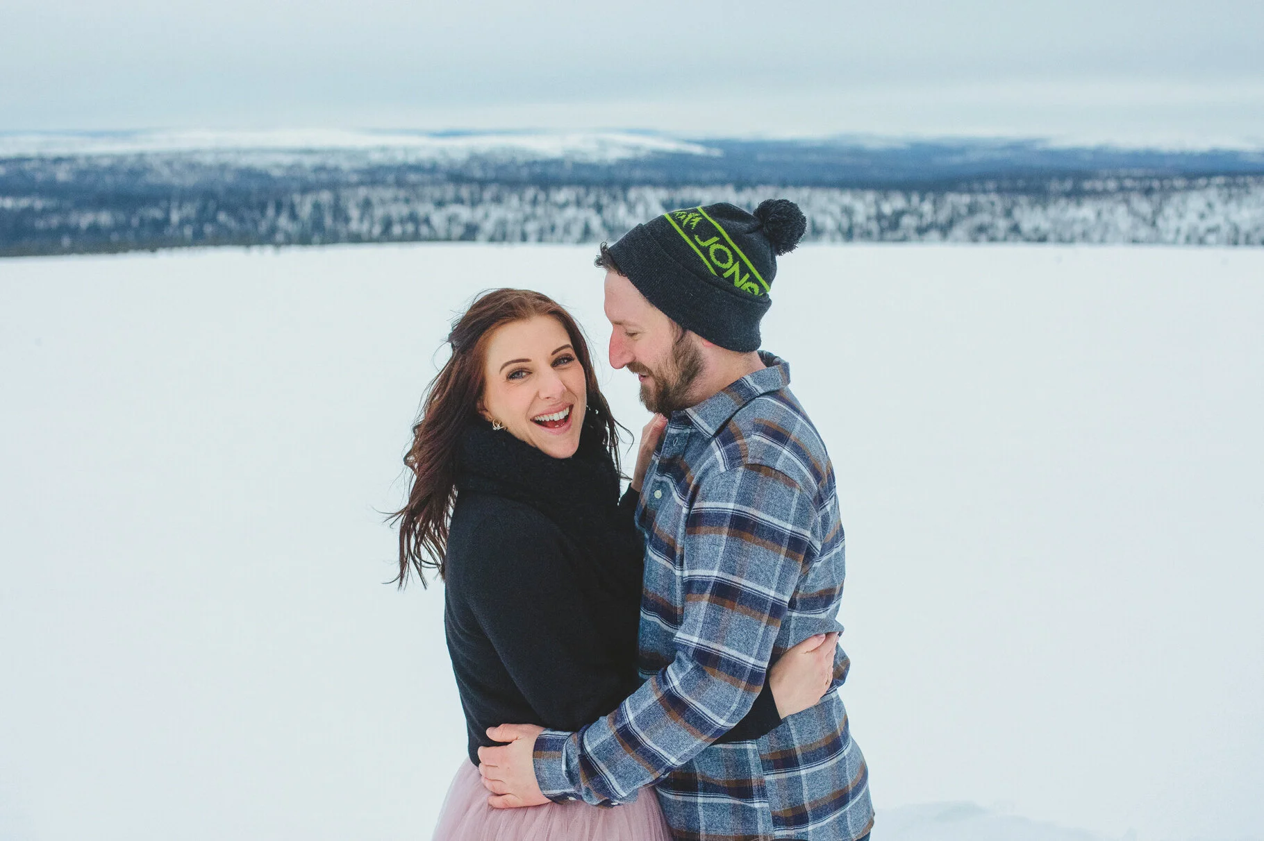 Elopement photos Lapland. A smiling woman and man embrace in a snowy landscape, with snow-covered trees and mountains in the background.