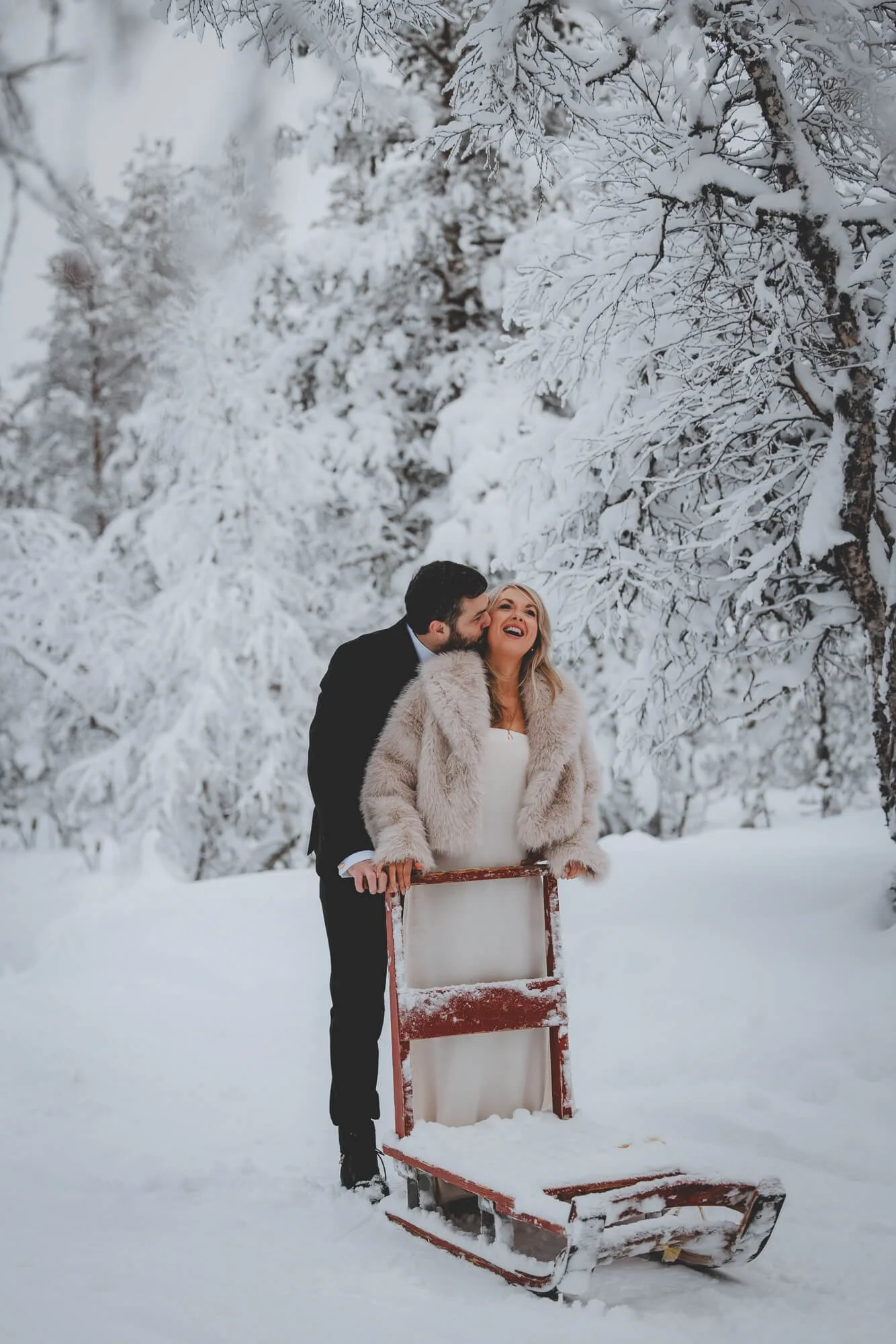Wedding photographer Rovaniemi and Lapland. A couple standing outdoors in a snow-covered landscape, with the man kissing the woman's cheek while she smiles, behind a wooden sled.