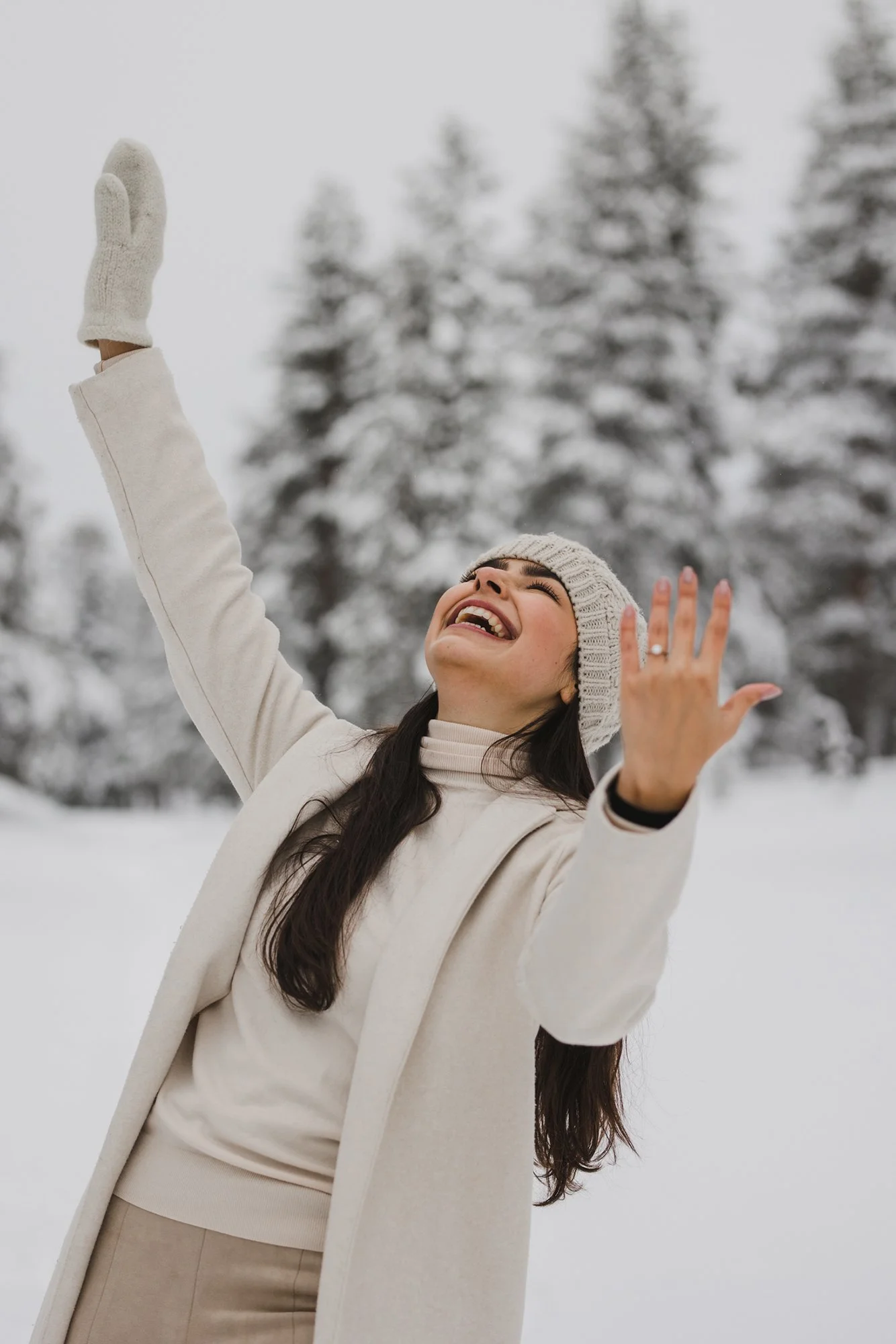 A woman smiling and raising her arms in a snowy outdoor setting with snow-covered trees in the background.