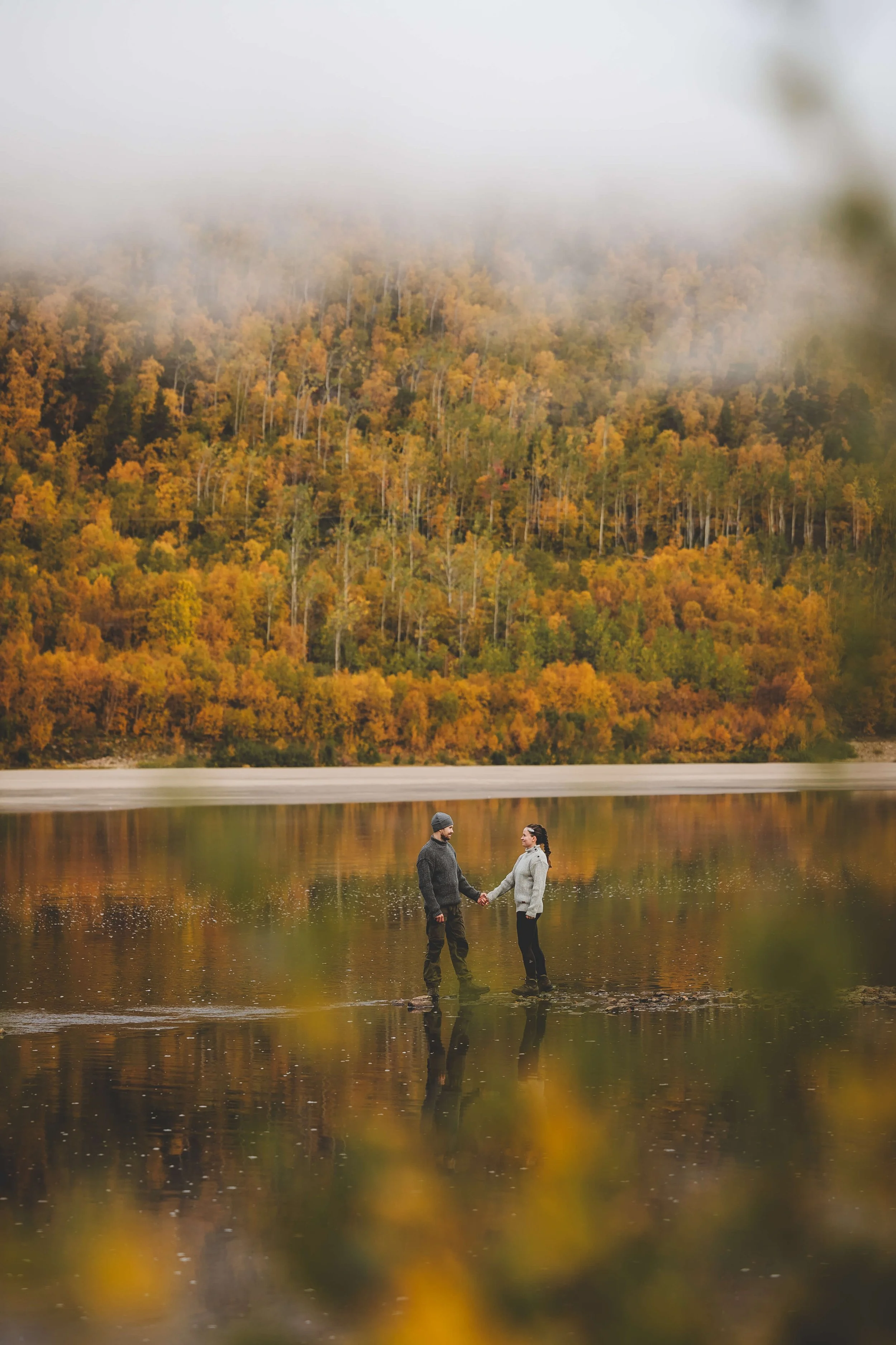 A couple holding hands and standing in a calm lake with a backdrop of colorful autumn trees and fog on a mountain.