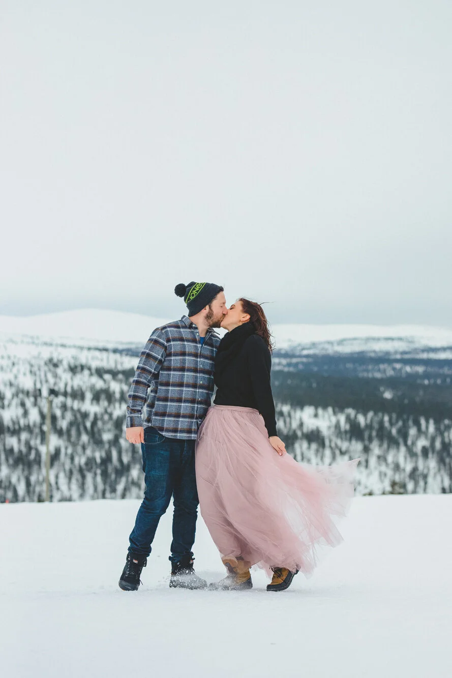 A couple sharing a kiss in a snowy landscape, with snow-covered trees and mountains in the background. Elopement in Lapland.
