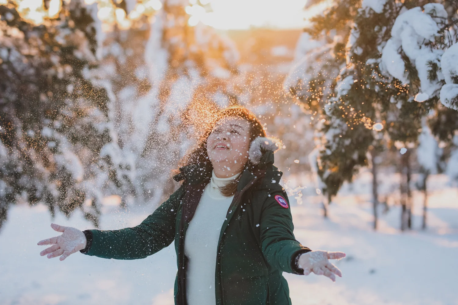 A girl in a winter coat with fur trim smiling and playing in falling snow during sunset surrounded by snow-covered trees.