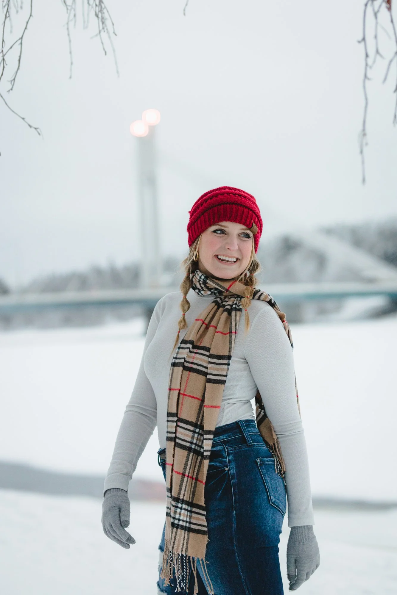 A young woman in a red knit hat, plaid scarf, white long sleeve shirt, and blue jeans standing outdoors in a snowy landscape with trees and a bridge, smiling and looking to the side.