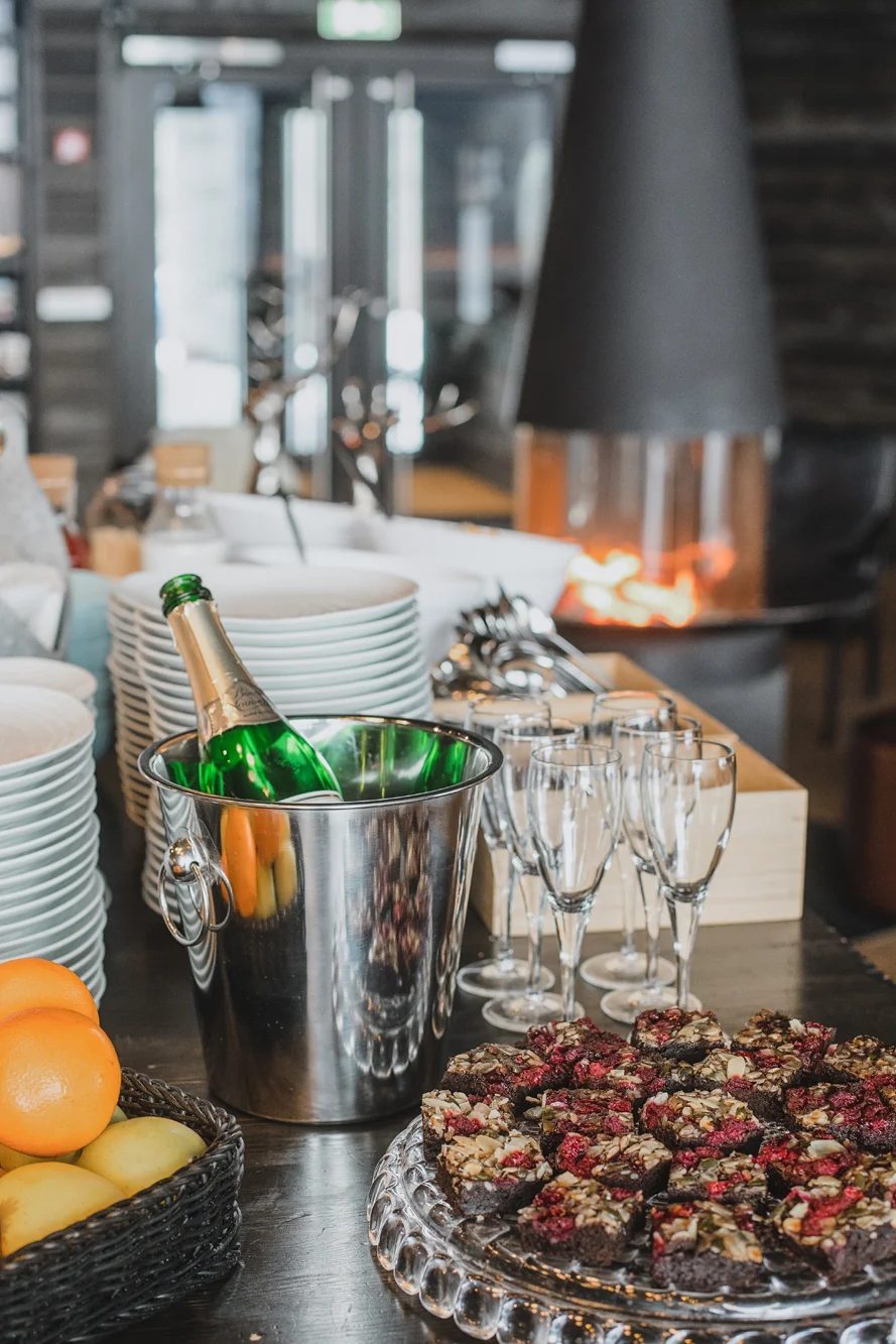 A photo of A table set for a celebration with a bottle of champagne in an ice bucket, empty champagne glasses, a tray of chocolate brownies topped with nuts and berries, a basket of oranges and lemons, and a background fireplace.