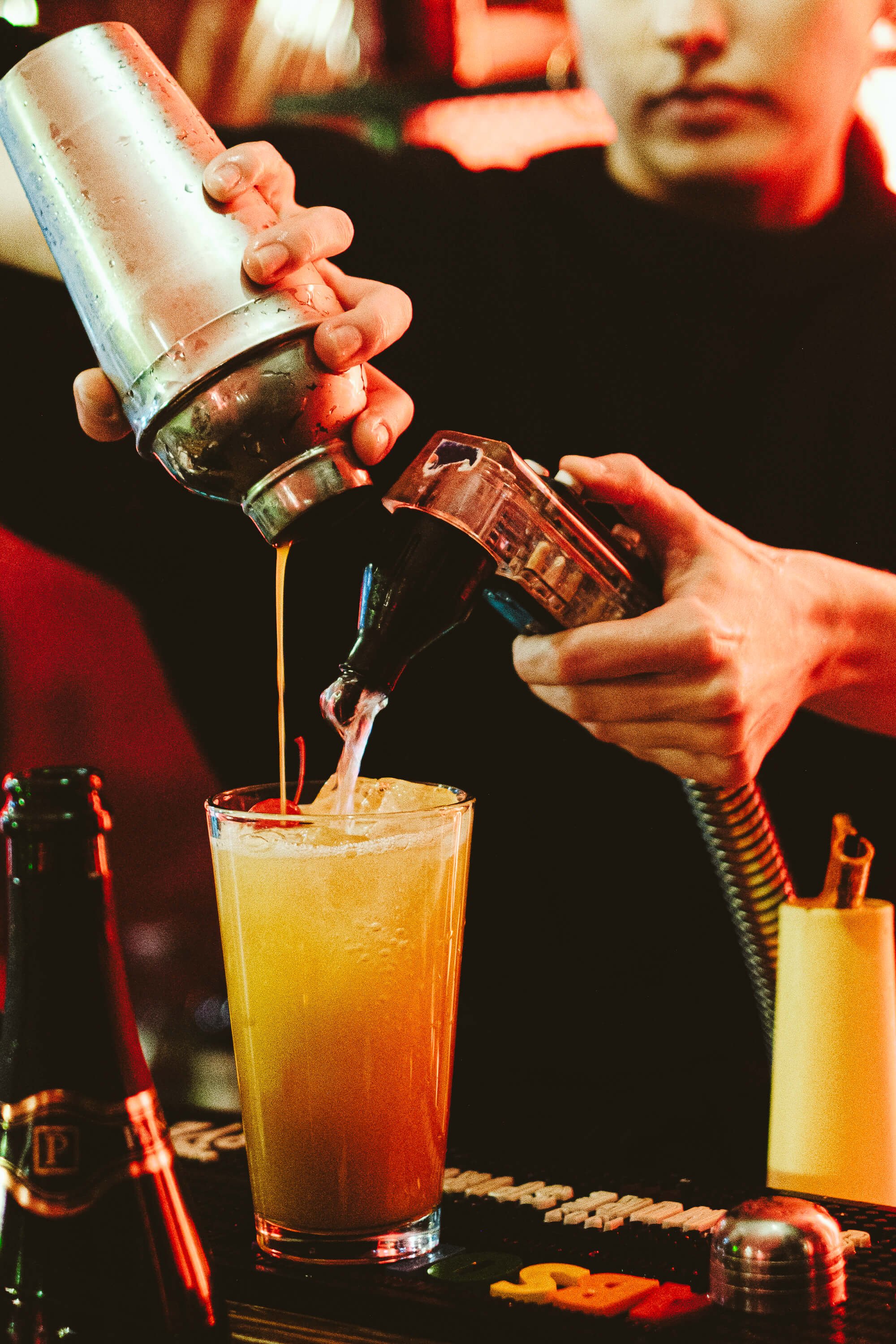 Bartender pouring a cocktail into a glass from a shaker with a soda gun, in a bar setting.