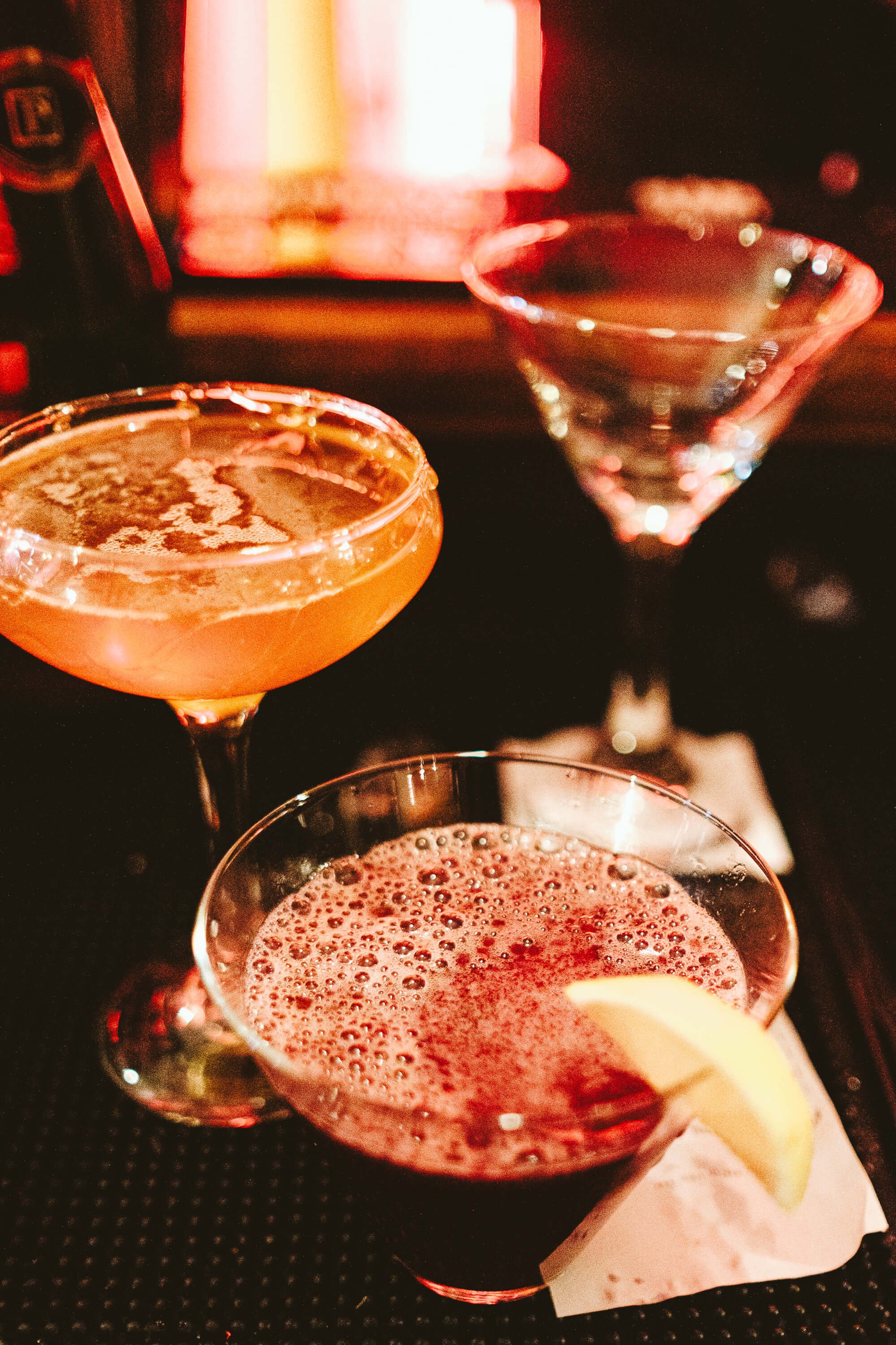 Three cocktails placed on a bar counter; a coupe glass with an orange drink, a martini glass with a clear liquid, and a rocks glass with a red drink garnished with a lemon slice.