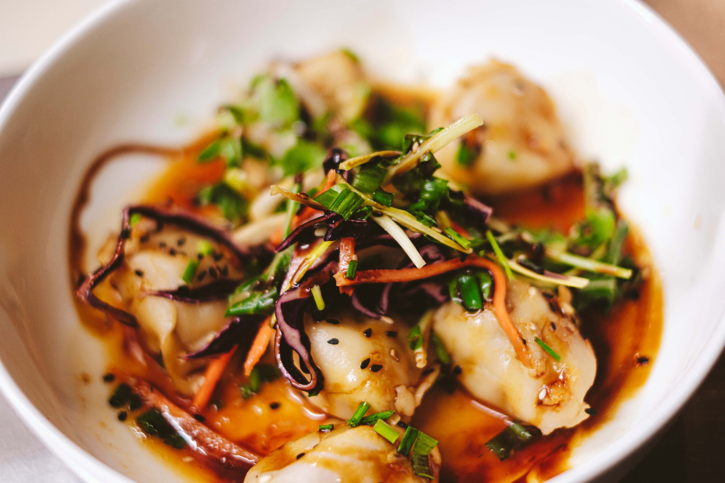 Dumplings in a bowl with sauce, garnished with sliced vegetables and herbs.