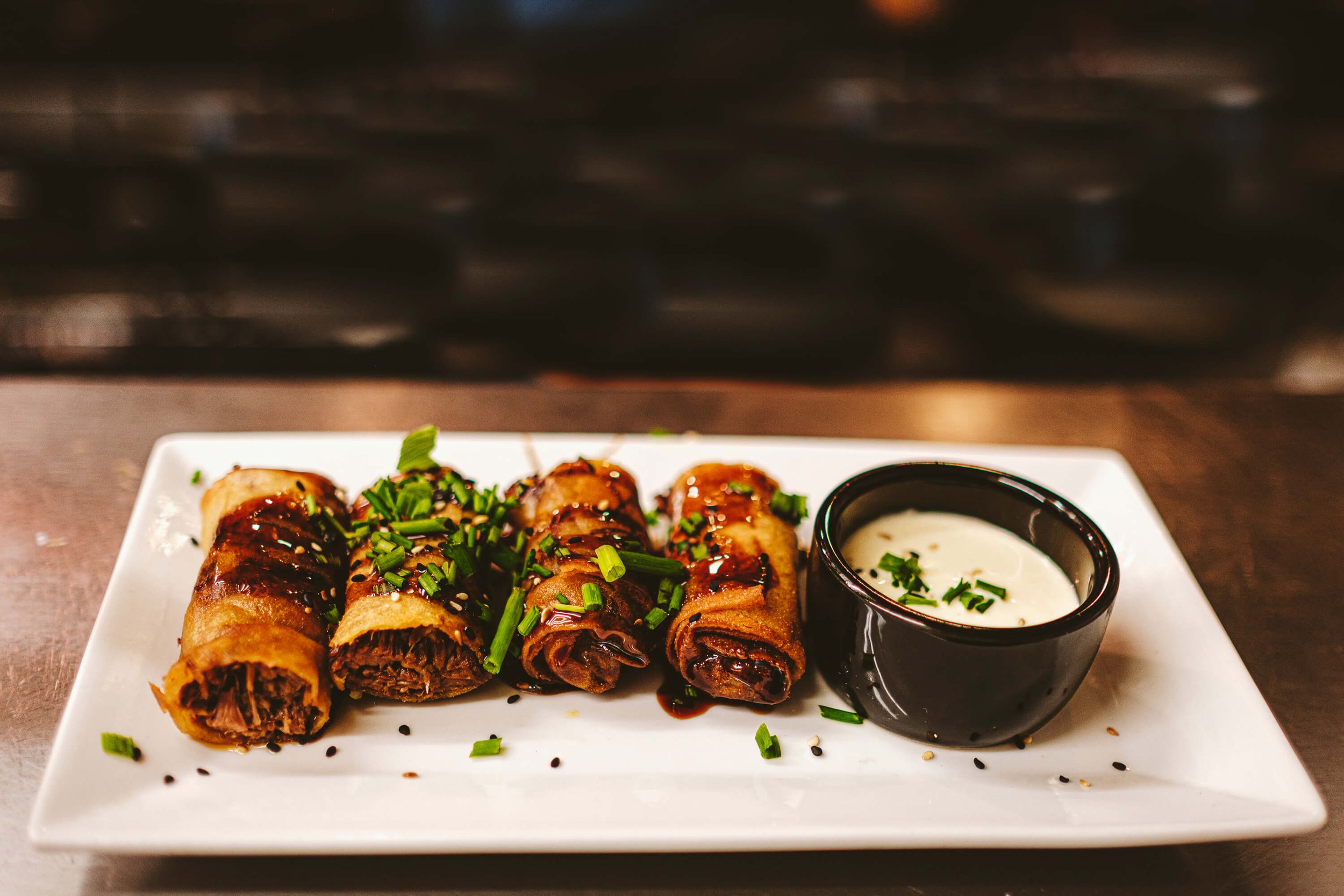 Crispy spring rolls garnished with green onions, sesame seeds, and sauce on a white plate with a side of dipping sauce in a small black bowl.
