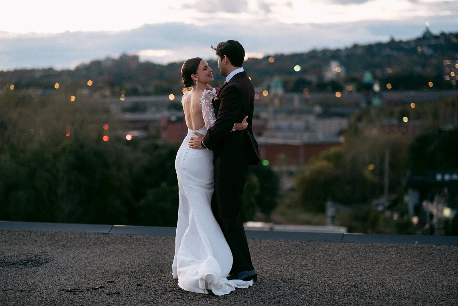 A bride and groom dancing outdoors at dusk, with a cityscape and hills in the background.