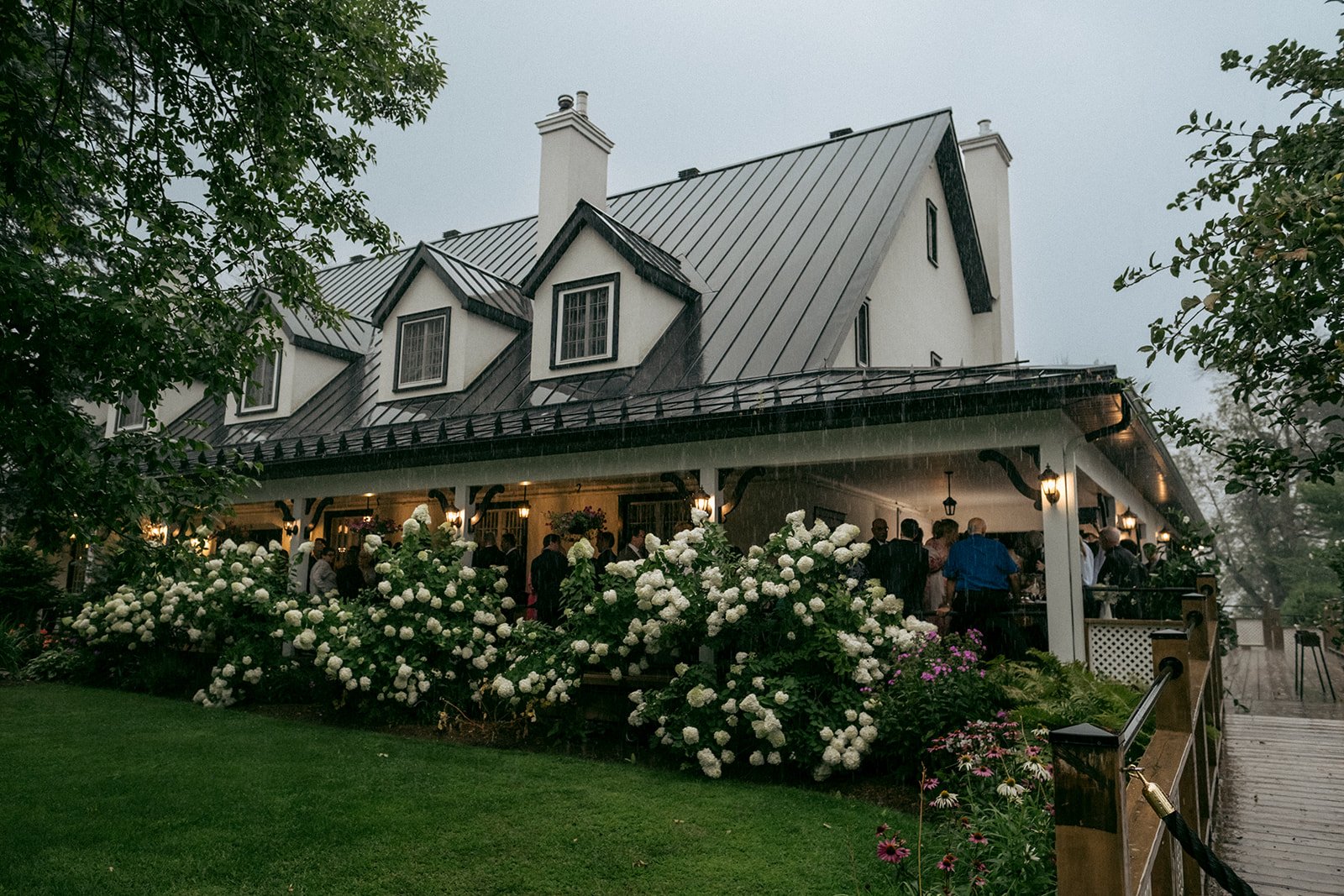 A large white house with a steep metal roof and multiple dormer windows, bustling with people on a covered porch during a rain shower, surrounded by lush green plants and white hydrangeas.