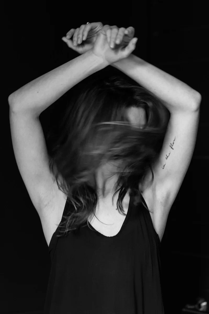 Black and white photo of a woman with long hair, wearing a black dress, with her head turned down and hair covering her face, arms crossed above her head, against a dark background.