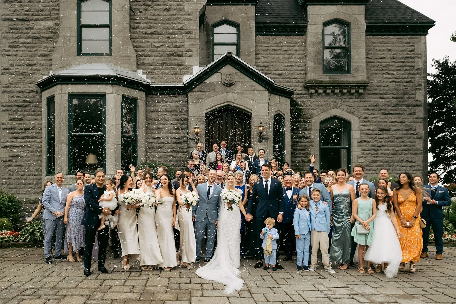 Large group of wedding guests, including bride and groom, people of various ages, dressed in formal attire, gathered on steps outside a stone building, celebrating and throwing confetti.