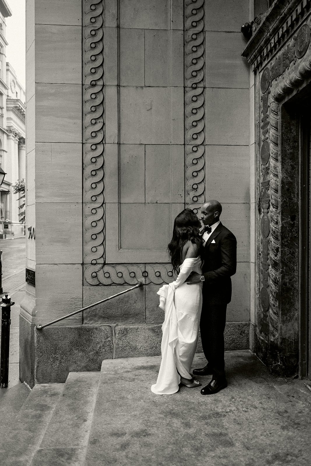 A couple dressed in formal wedding attire standing close and embracing outside a building with ornate decoration, on steps leading up to a doorway.