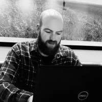 A bearded man with a checked shirt sitting at a desk, working on a laptop.