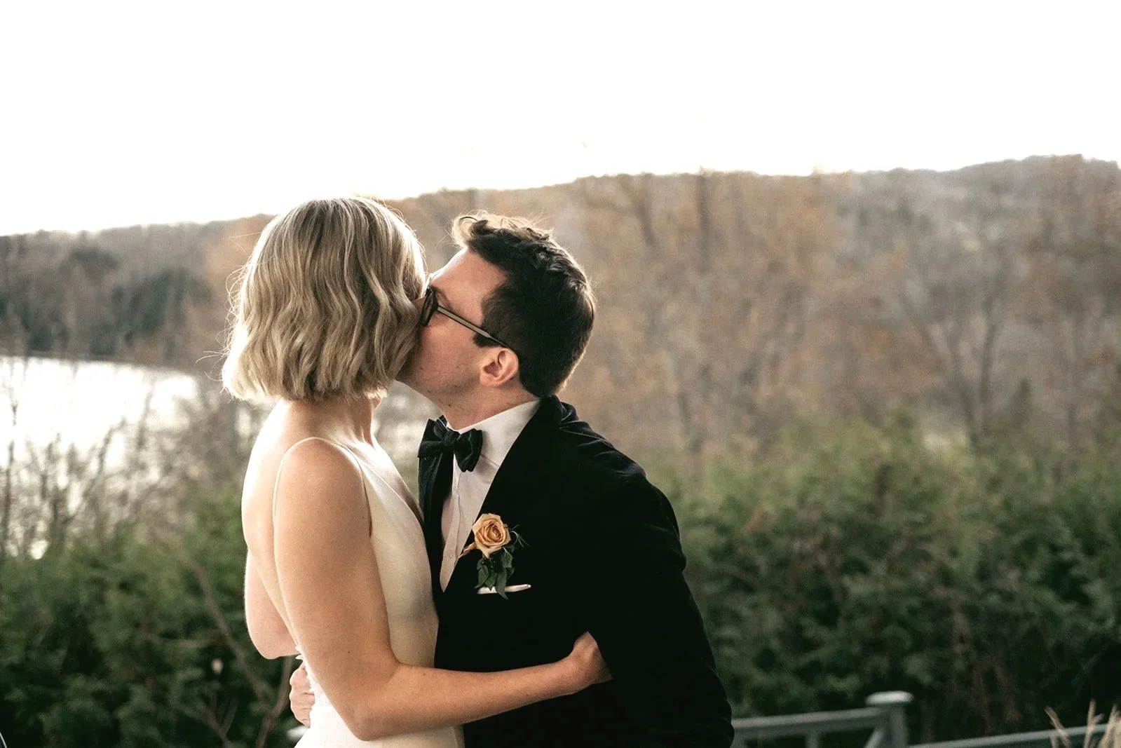 A couple dressed in formal attire sharing a kiss outdoors, with trees and hills in the background.
