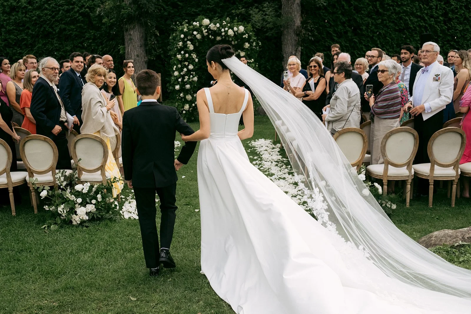 Bride and a young boy holding hands walking down an outdoor wedding aisle lined with chairs and guests, with an arch of white flowers and green trees in the background.