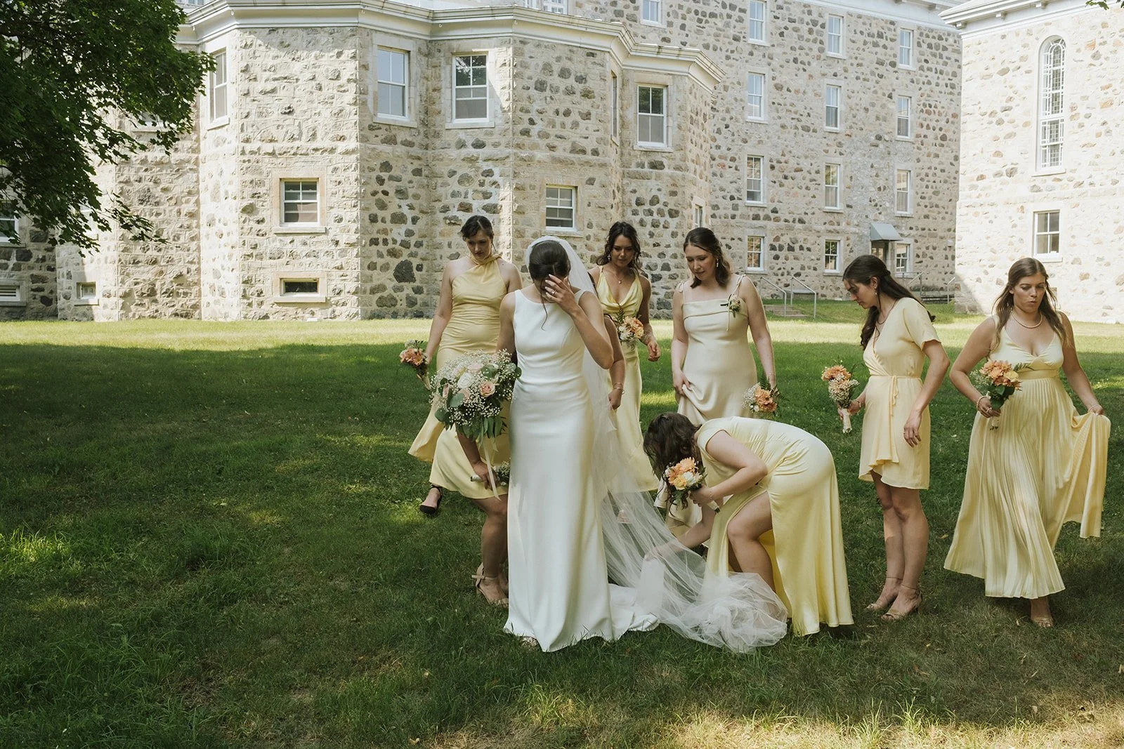 A bride in a white wedding gown and veil is surrounded by bridesmaids in yellow dresses outside on grass, with a large stone building in the background. Some bridesmaids are holding bouquets.