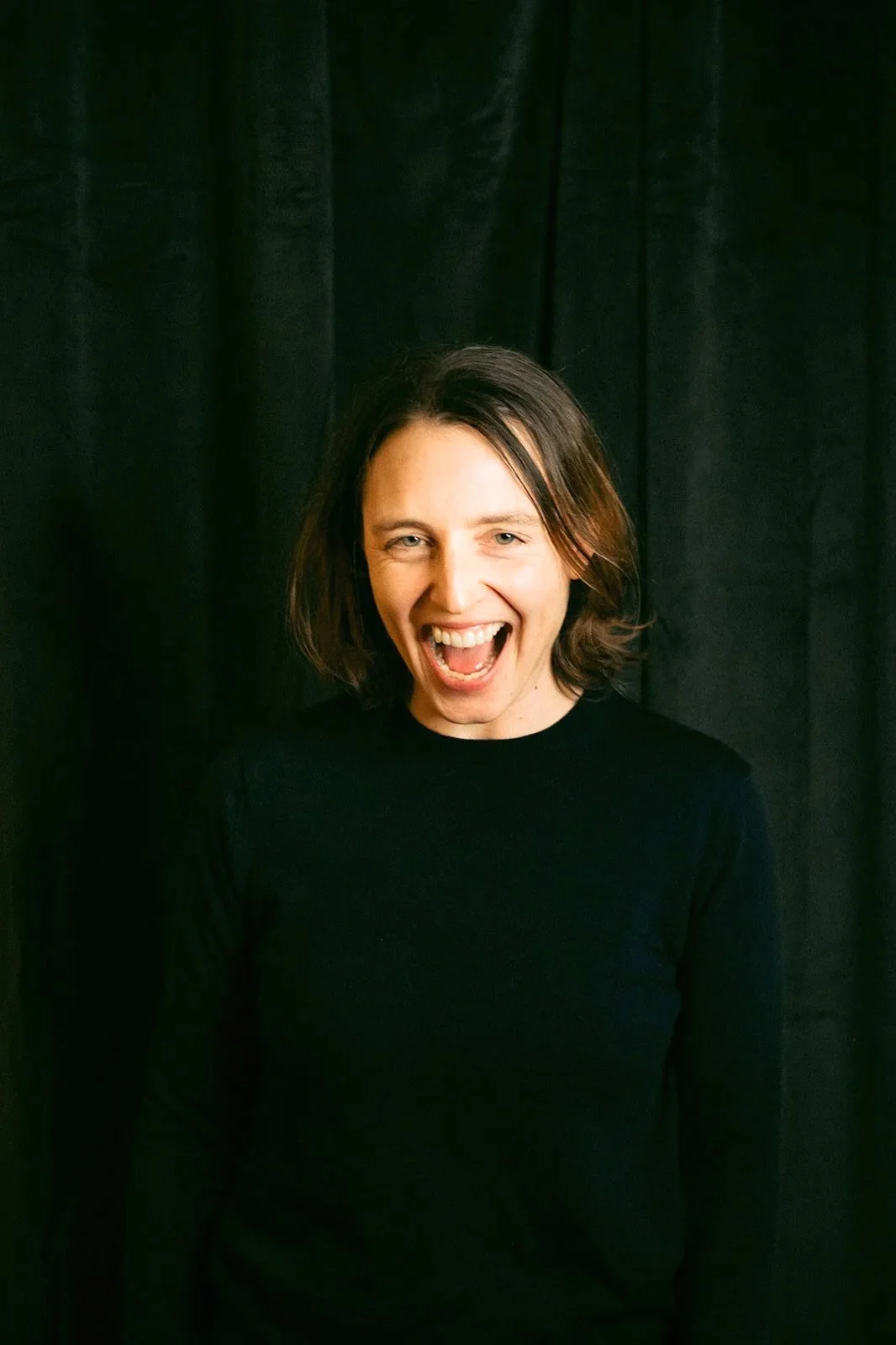 A woman with shoulder-length brown hair, wearing a black long-sleeve shirt, is smiling and laughing in front of a dark curtain background.