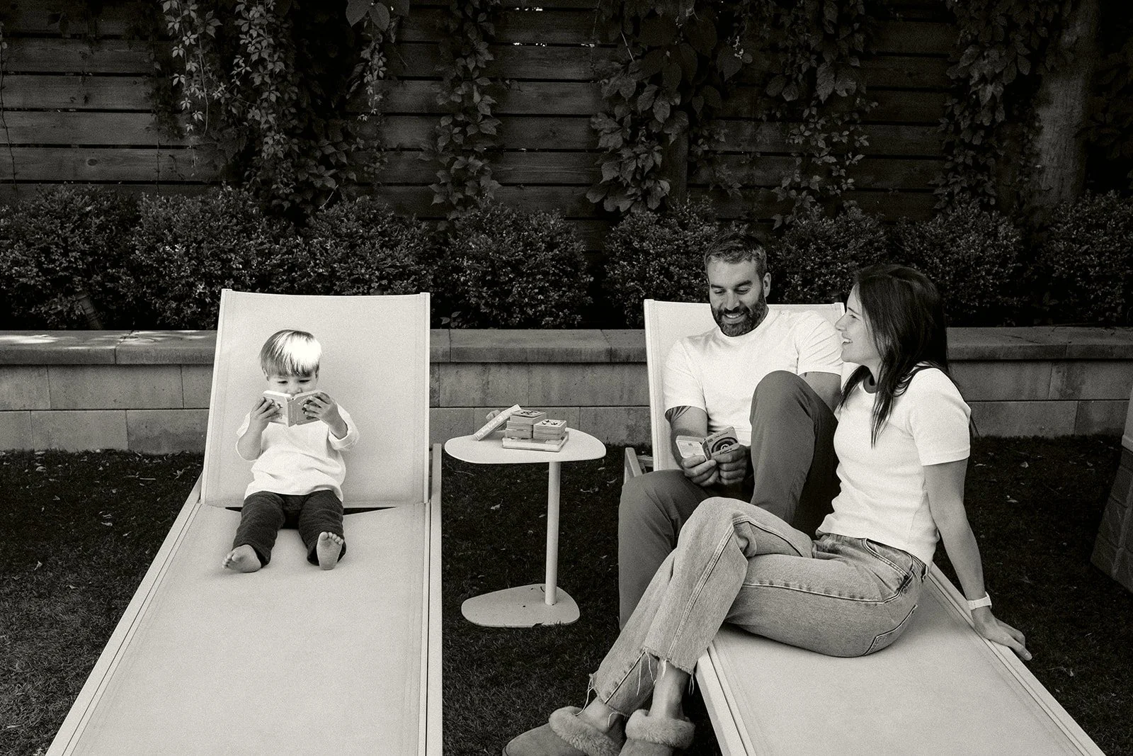 A family of three relaxing in a backyard, with a woman and man sitting on lounge chairs and a young boy reading a book on a separate lounge chair. There is a small round table between them with books or magazines on it, and a wooden fence with bushes and trees in the background.
