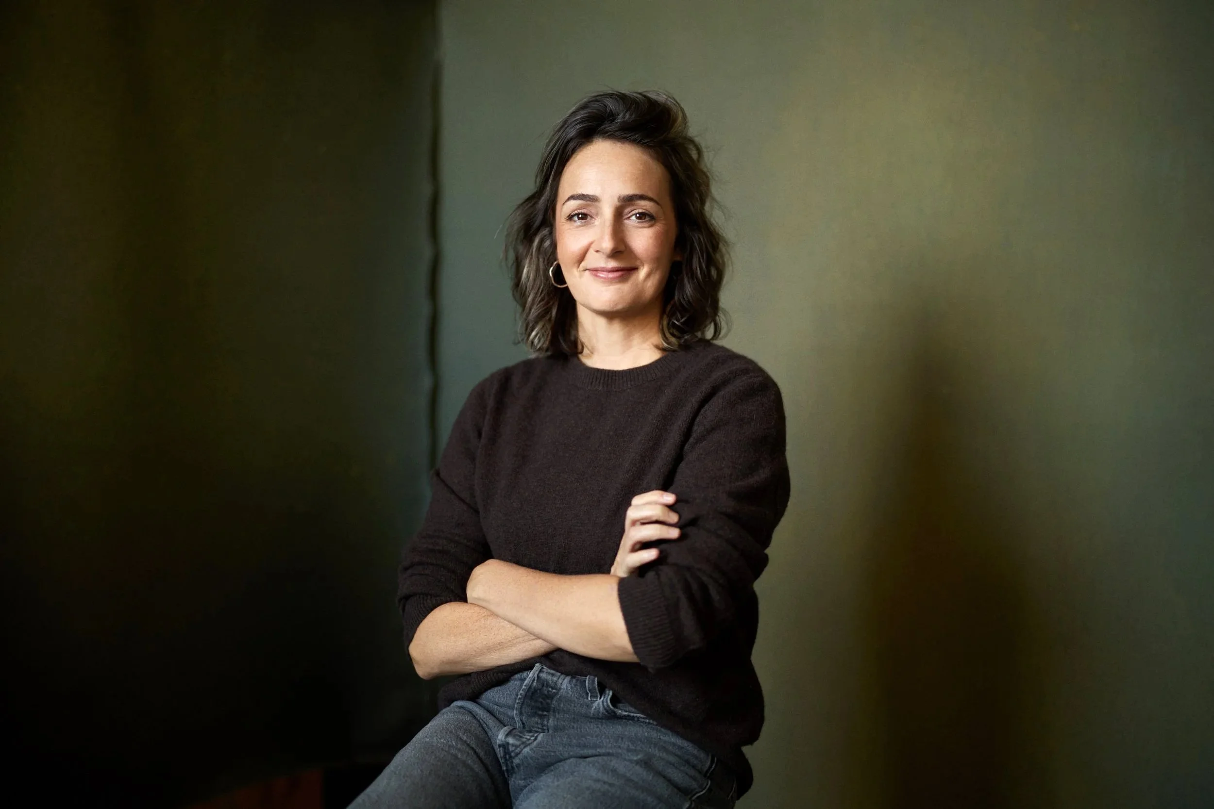A woman with shoulder-length dark hair, wearing a black sweater and gray pants, sitting with her arms crossed against a greenish wall, smiling at the camera.