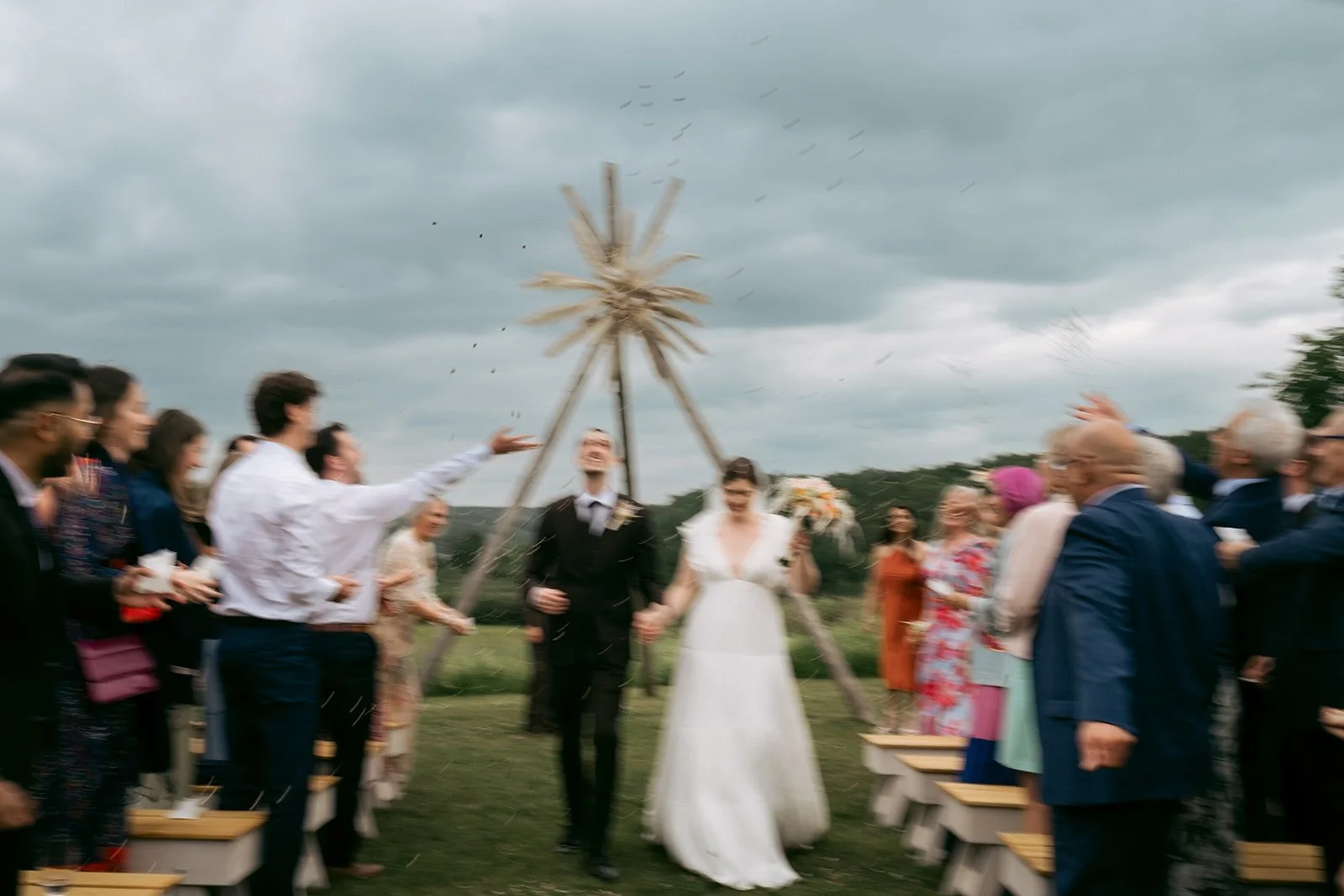 A wedding celebration taking place outdoors, with the bride and groom holding hands as guests throw rice or flower petals in the air.