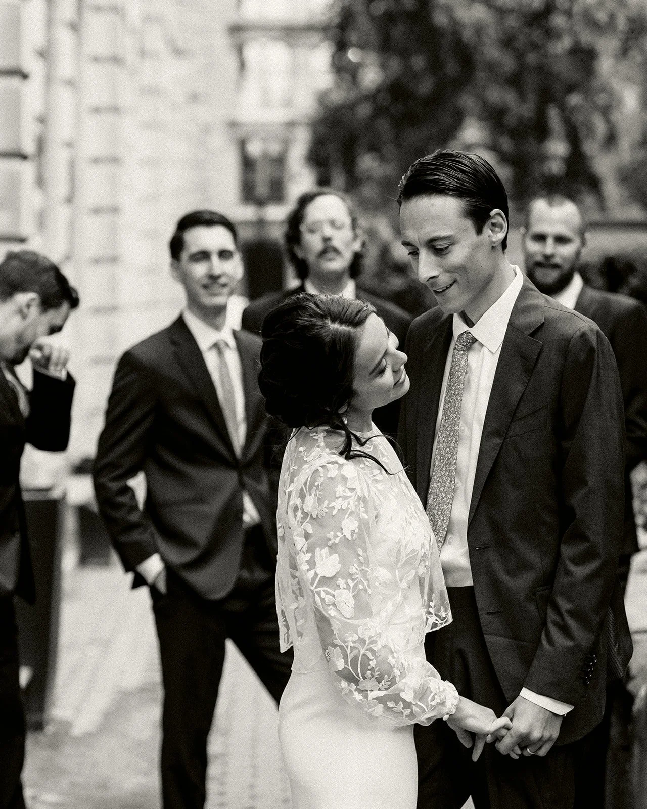 Black and white photo of a bride and groom holding hands, looking at each other, with three men in suits in the background.