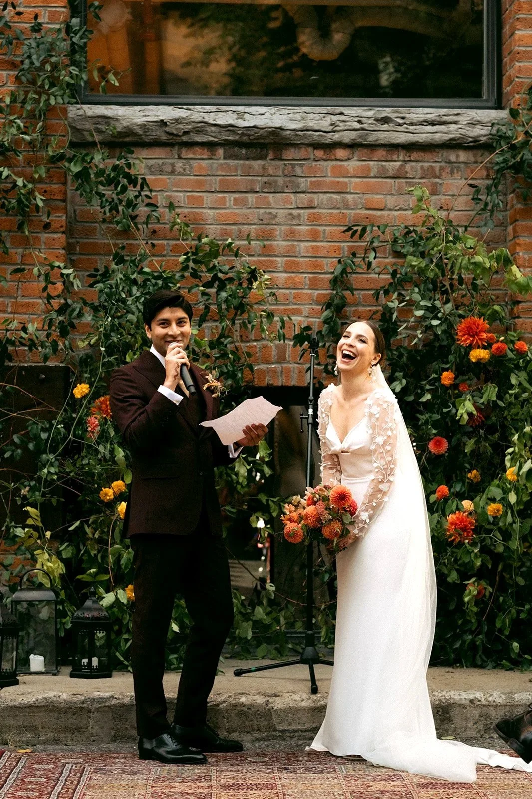 A wedding ceremony with a groom and bride standing in front of a brick wall and flower arrangement. The groom, dressed in a dark suit, is holding a microphone and a paper, smiling. The bride, in a white wedding gown with floral details and holding a bouquet of red and orange flowers, is laughing. There are lanterns on the ground, and the scene appears festive and joyful.