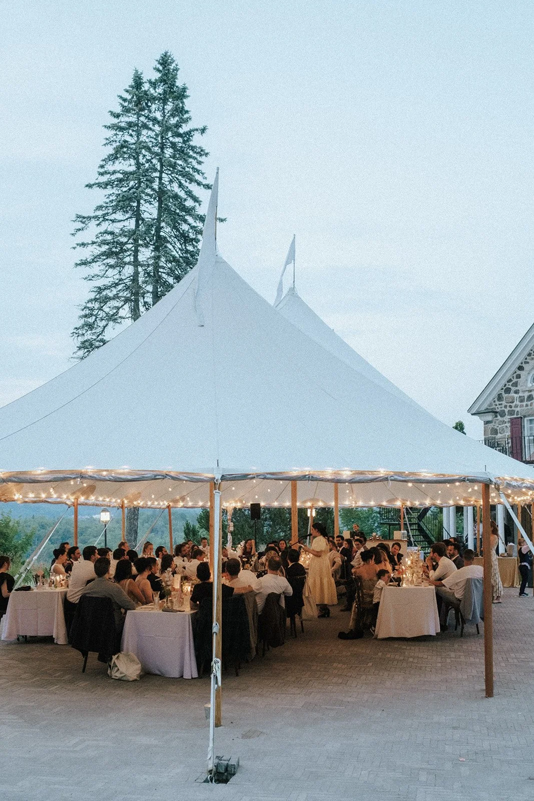 An outdoor event under a large white tent with string lights, with many people seated at tables, a woman standing and speaking, a stone building to the right, and tall trees in the background.