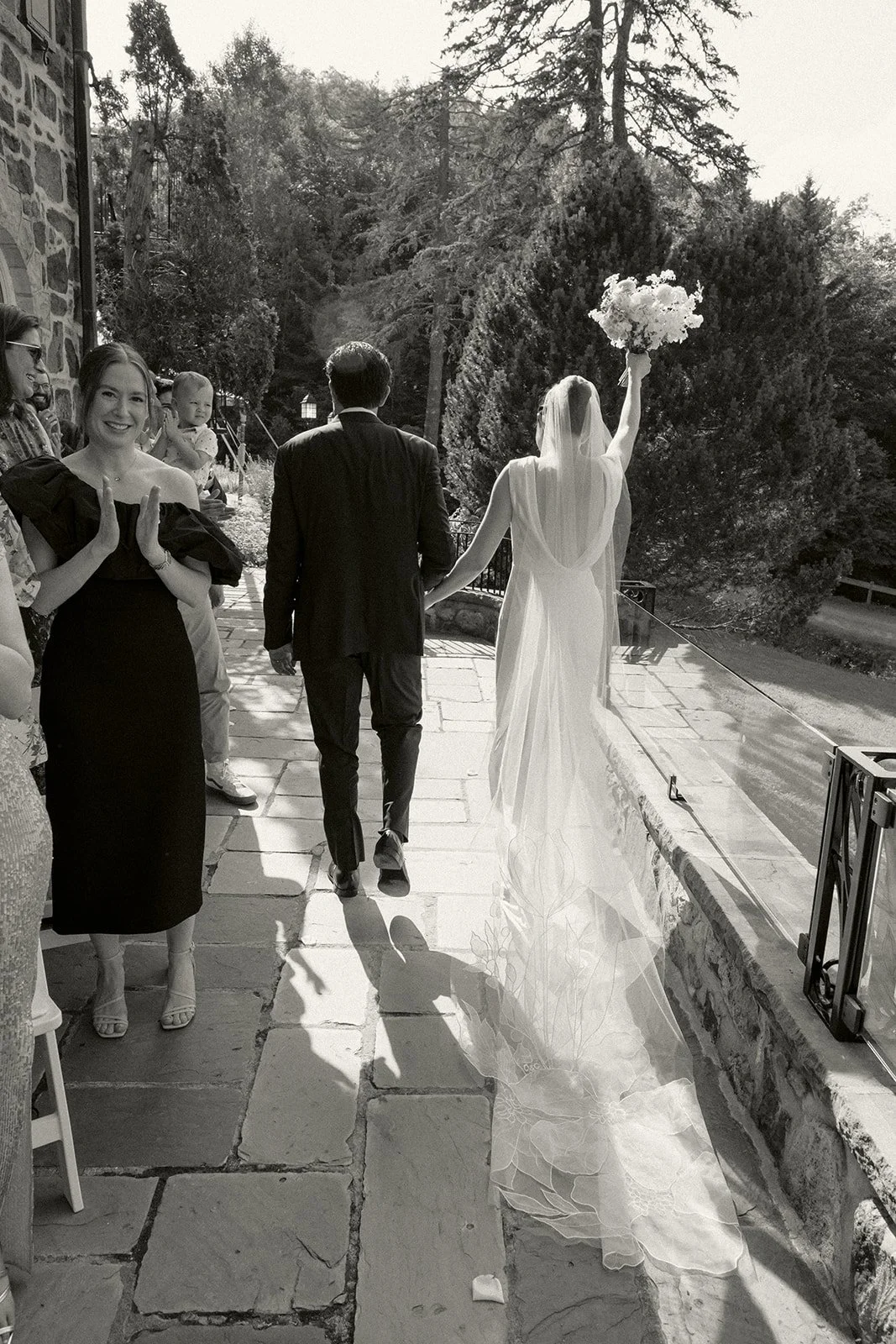 Bride and groom walking hand in hand outside after wedding ceremony, with wedding guests on the side, in black and white.