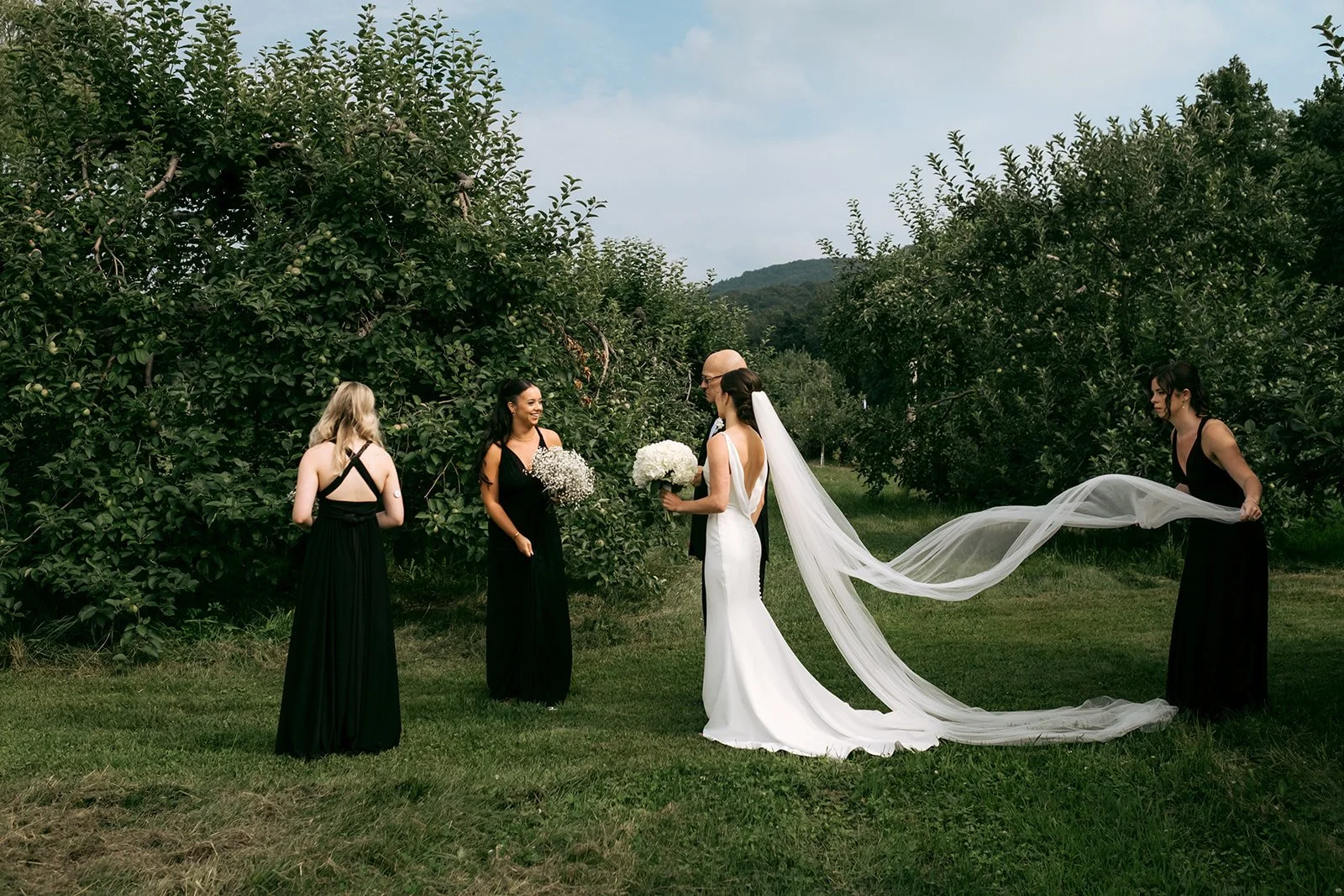 A wedding ceremony outdoors in a green orchard with five women: a bride in a white gown with a long train and veil, holding a bouquet of white flowers, talking to another woman in a black dress, with three bridesmaids in black dresses standing nearby, one of whom is adjusting the bride's train.