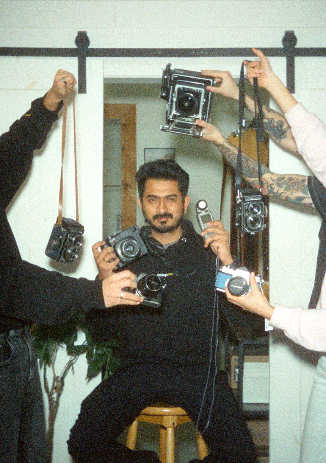 A man with dark hair and tattoos is sitting on a stool, surrounded by multiple cameras and photographic equipment held by others.