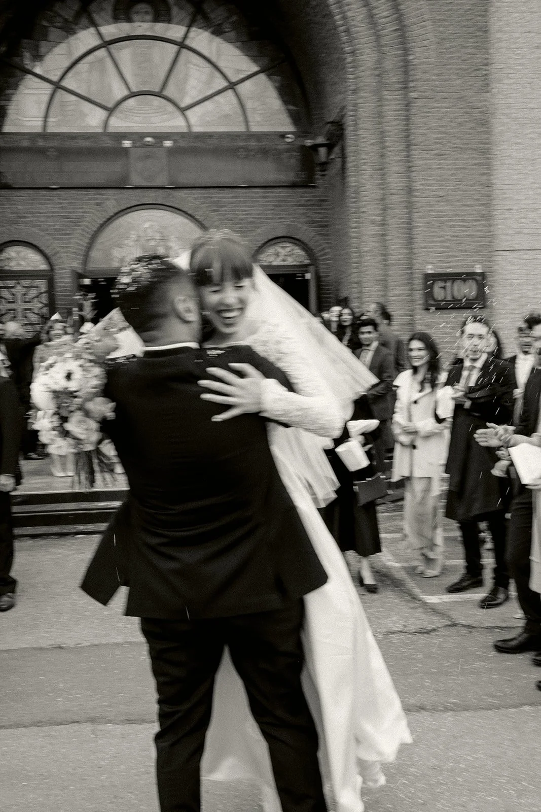A couple in wedding attire dancing and celebrating outside a building, with onlookers watching and clapping in the background.
