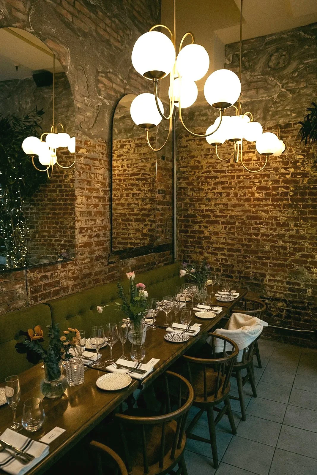 A cozy restaurant interior with a long wooden table set for dinner, flower arrangements, brick walls, a large mirror, green bench seating, wooden chairs, and modern chandelier lighting.