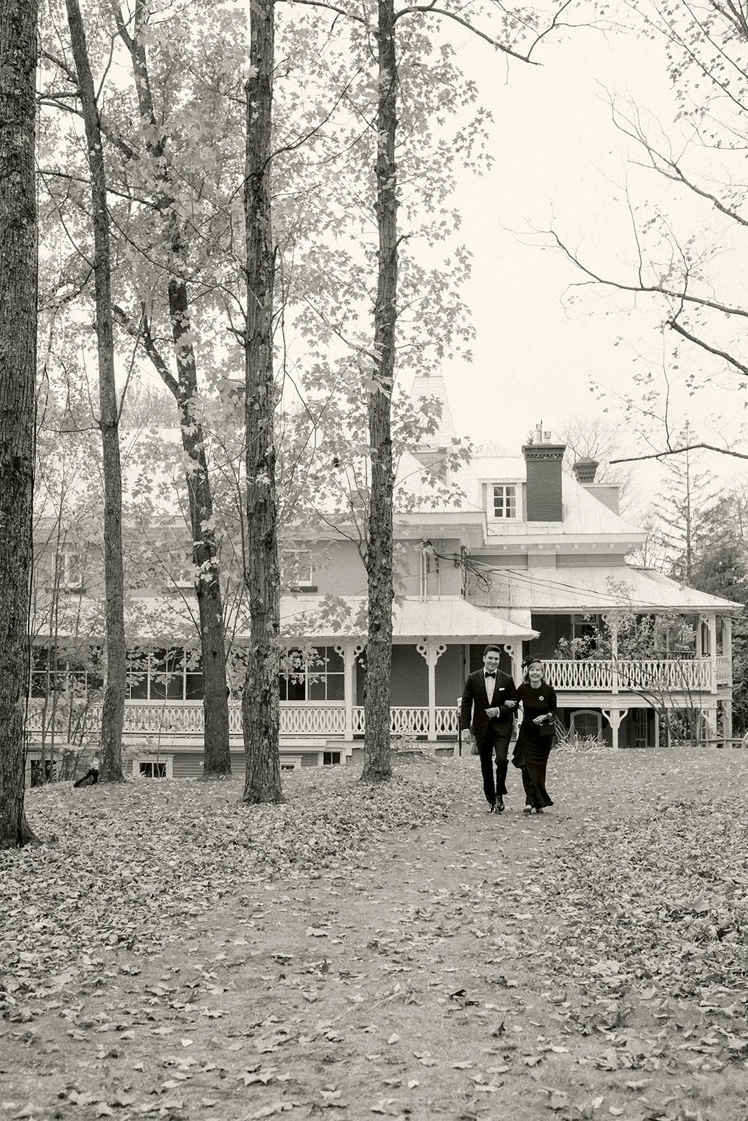 A man and woman in formal attire walking on a leaf-covered path in front of a large old house with a wraparound porch and a balcony, surrounded by trees.