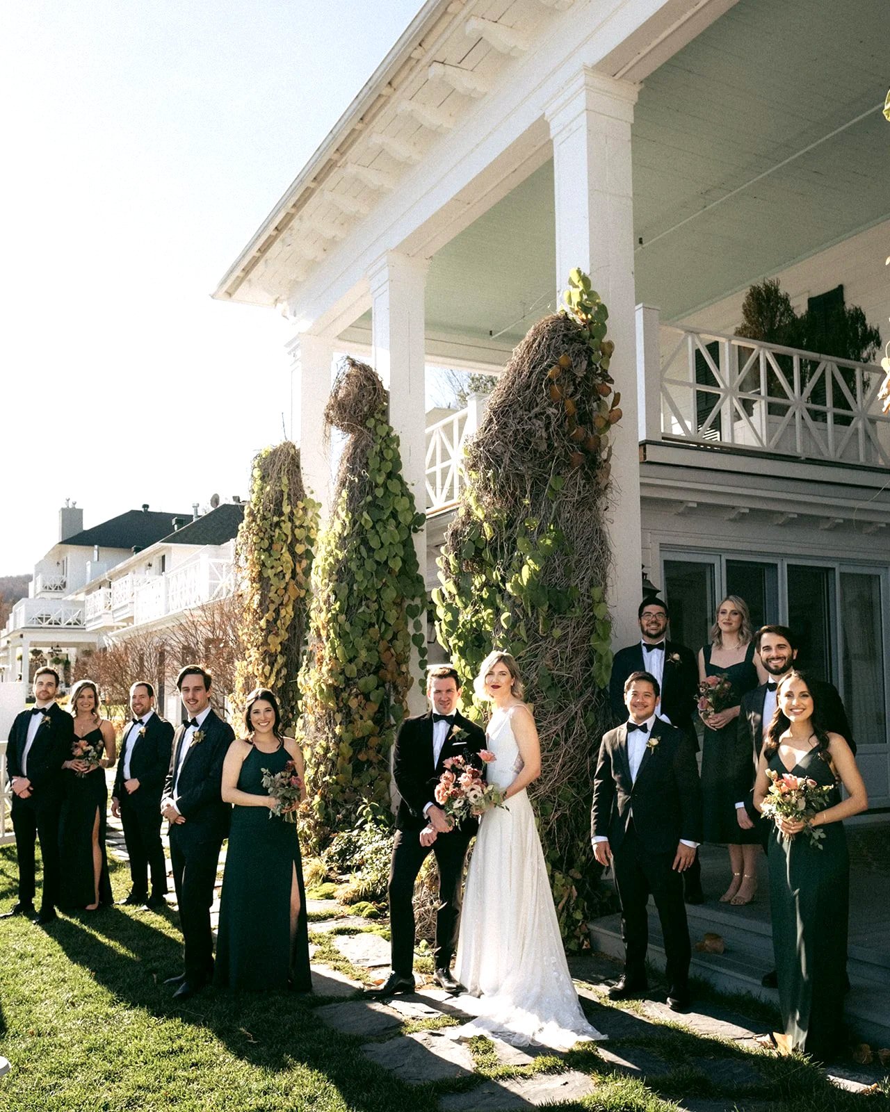 A group of wedding guests standing outside a white house with a porch, dressed in formal attire, with some women holding bouquets and men in tuxedos, on a sunny day.