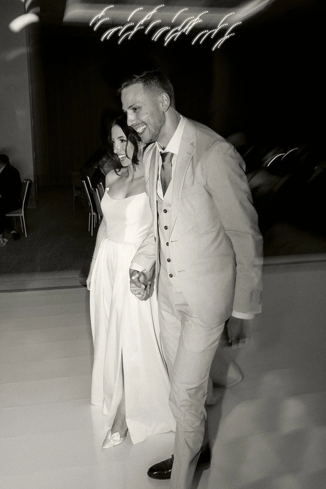 Black and white photo of a happy couple holding hands, dressed in formal attire, walking indoors with a blurred background.