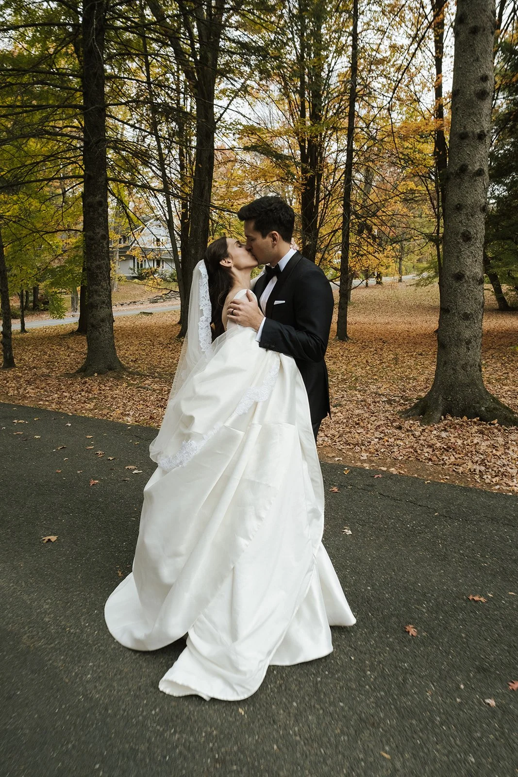 A bride and groom sharing a kiss, with the bride in a white wedding gown and veil, and the groom in a black tuxedo, standing on a paved path surrounded by autumn trees.