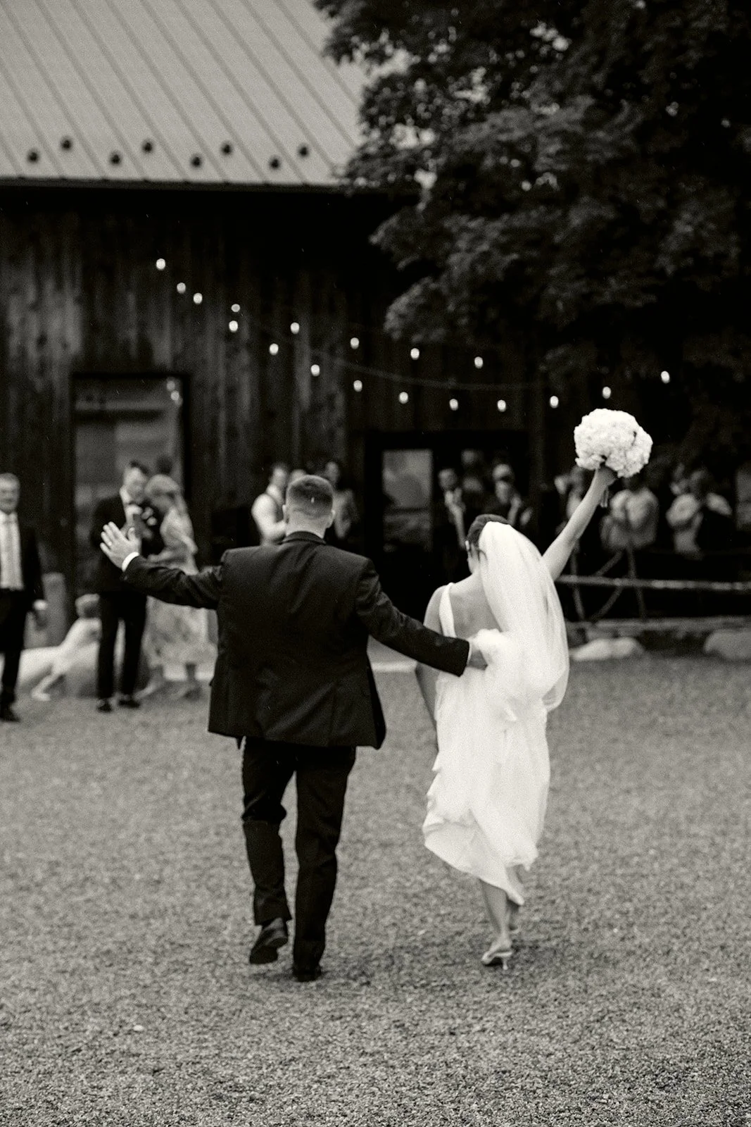 A black-and-white photo of a couple dancing outdoors, possibly at a wedding, with guests in the background. The woman is holding a bouquet and wearing a white dress, and the man is in a dark suit.