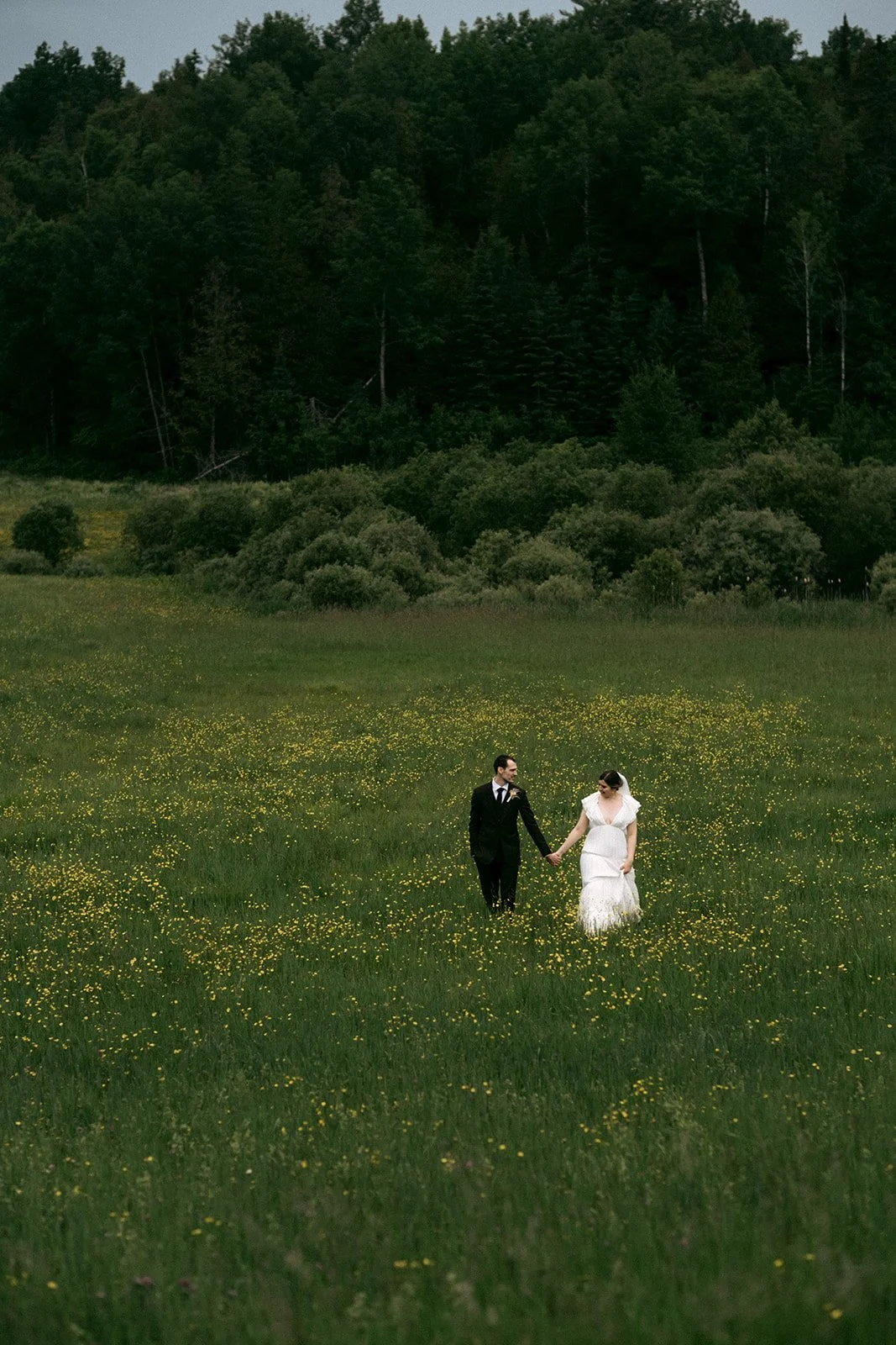 A bride and groom walking hand in hand across a field of yellow flowers, with dense green trees and a forested hillside in the background.