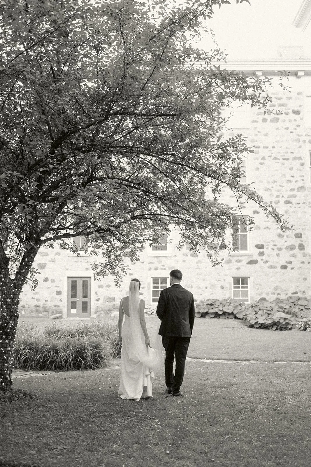 A couple in wedding attire walking together in a garden area with a tree and a stone building in the background.