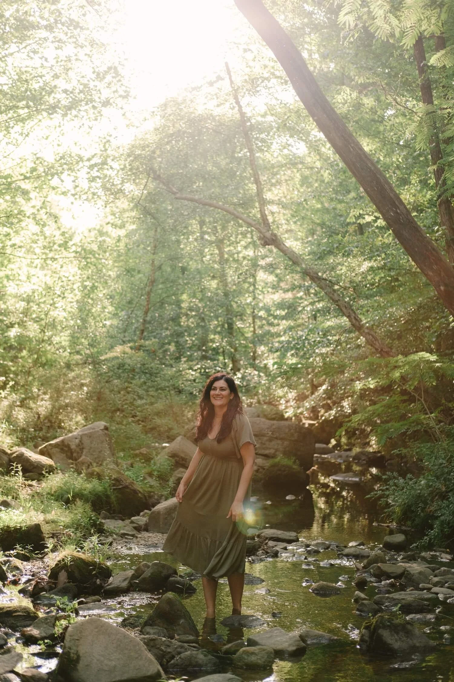 Kim Kimball smiles in a sun-drenched forest while wearing a green dress, embodying the essence of Life Transition Coaching.