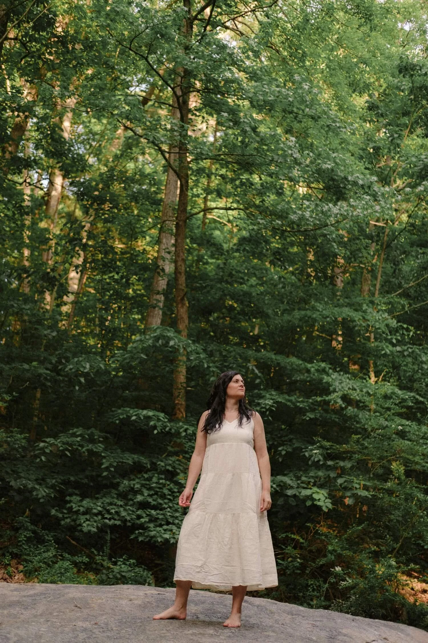 A woman named Kim Kimball stands on a large rock in the forest, representing the peace of Life Transition Coaching.