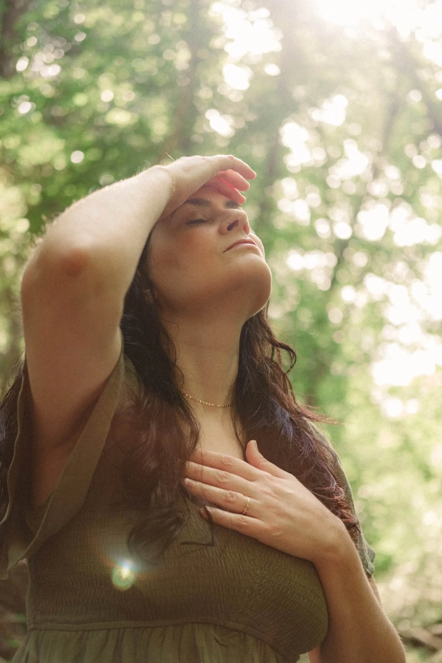 An embodiment coach, Kim Kimball, stands in a forest with eyes closed, hands on her head and chest in reflection.