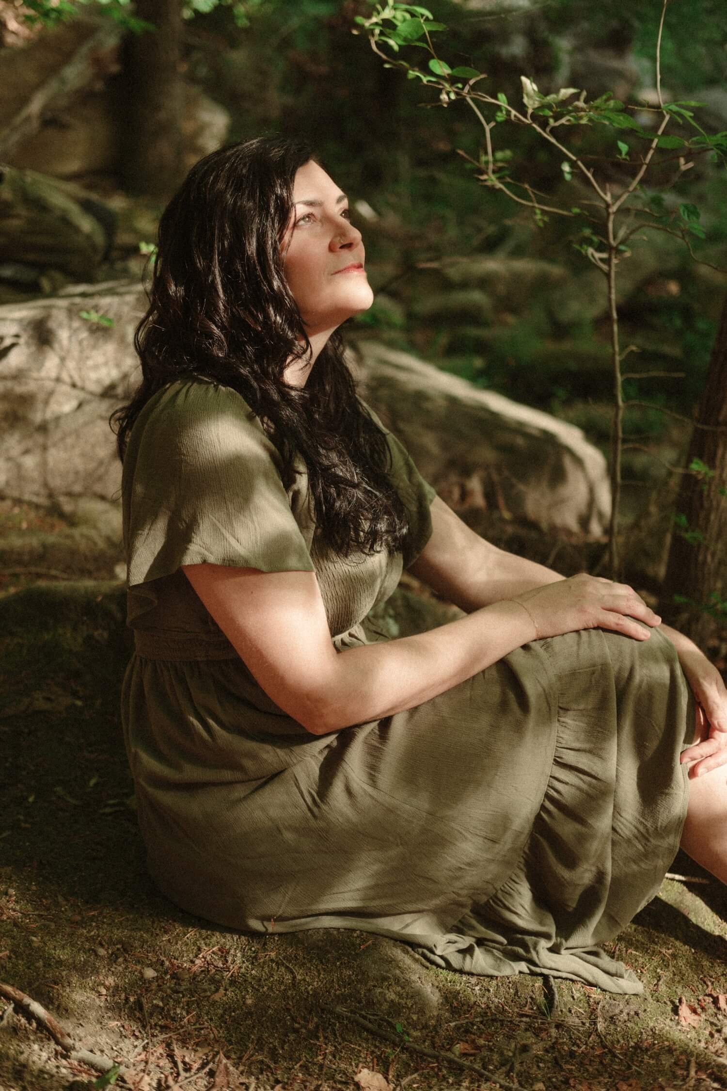 A woman in a green dress gazes up while resting on a rock in a wooded area, illustrating Life Transition Coaching.