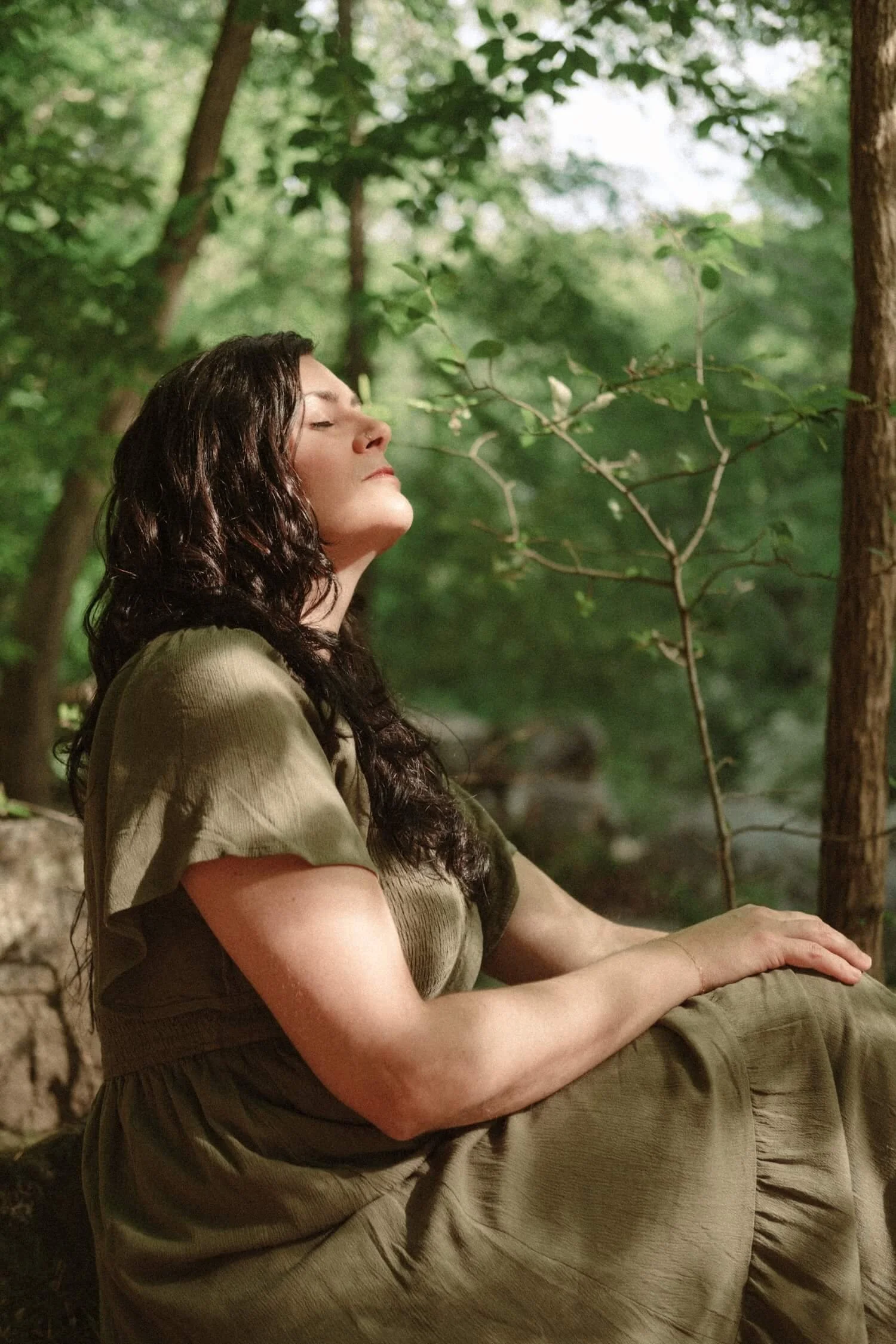 Kim Kimball, an embodiment coach, sits serenely on the forest floor with her eyes closed in the dappled light.